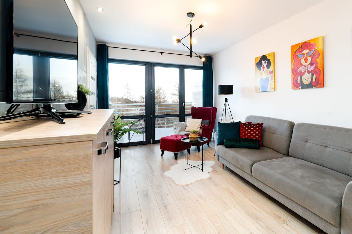 Un salón moderno con un sofá gris, un sillón rojo y una pequeña mesa redonda con una planta. Hay ventanas amplias y vistas al exterior.