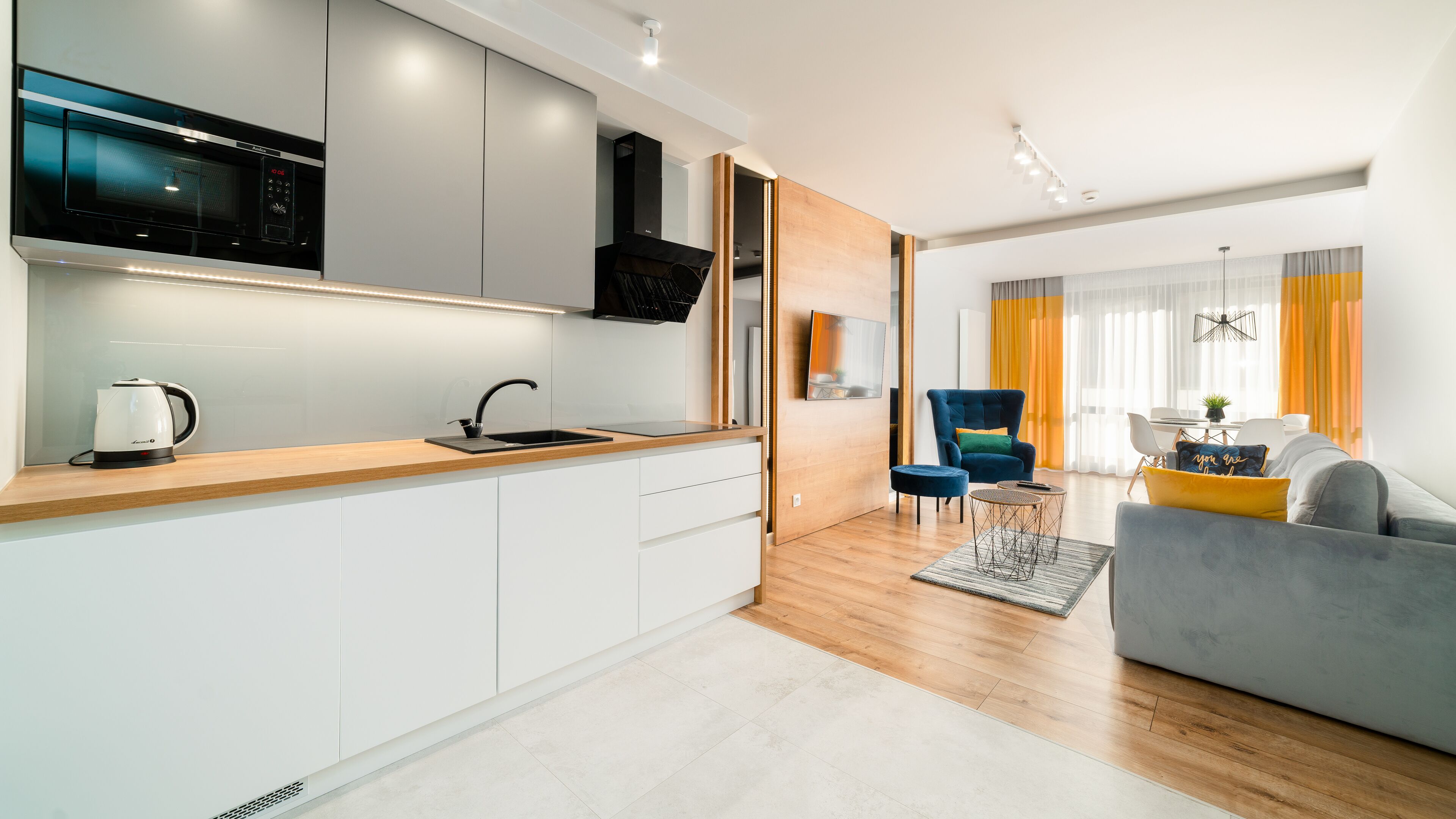 A modern kitchen with a microwave, kettle, and sink. There is a dining area with a sofa, chair, and table.