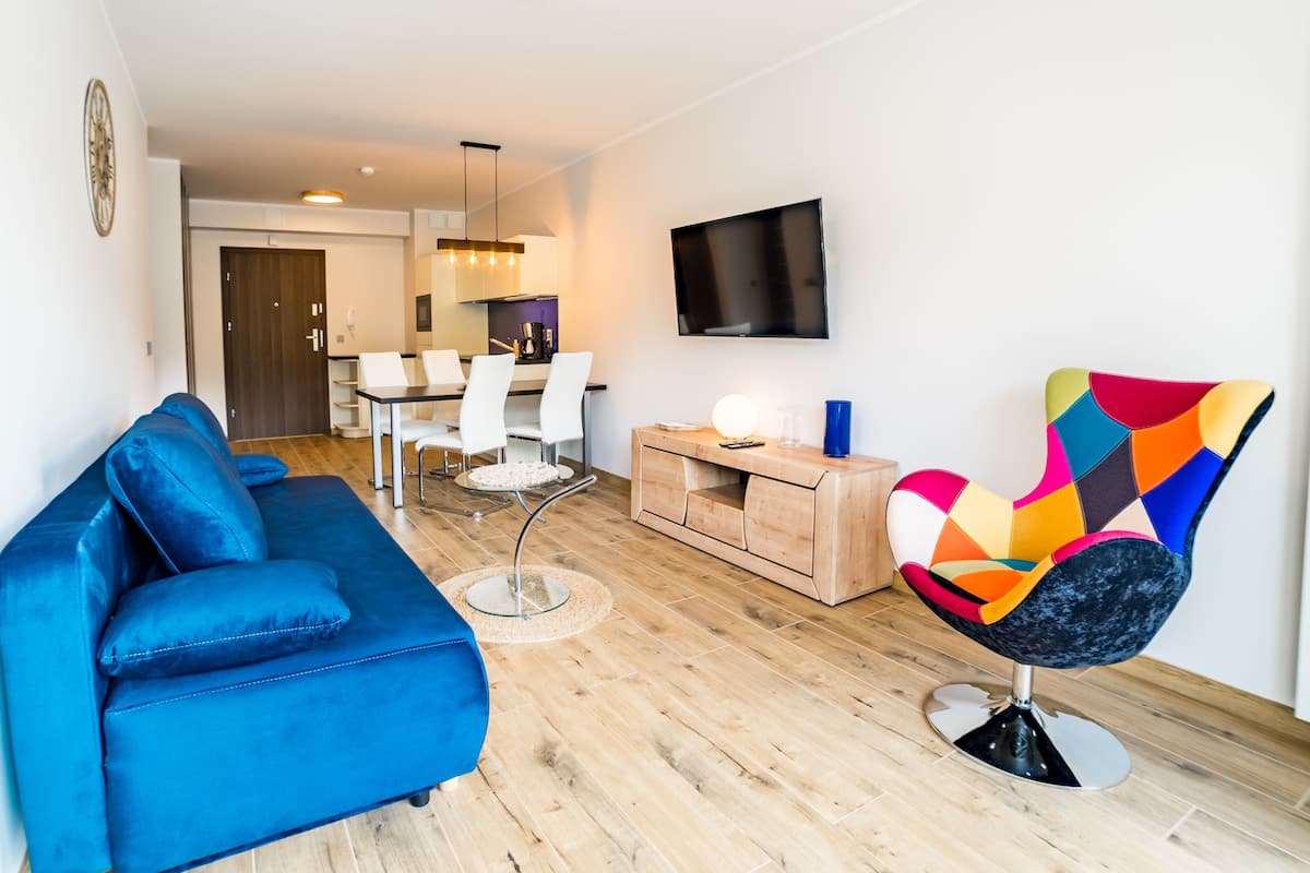 Una sala de estar moderna con un sofá azul, un sillón colorido, un televisor montado en la pared y un comedor con sillas blancas y una mesa de madera.