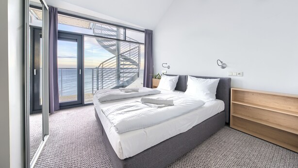 A modern hotel room with a large bed, a wooden headboard, a nightstand, and a view of the ocean through a sliding glass door. - Sun & Snow Triventi Mountain Residence (Karpacz)
