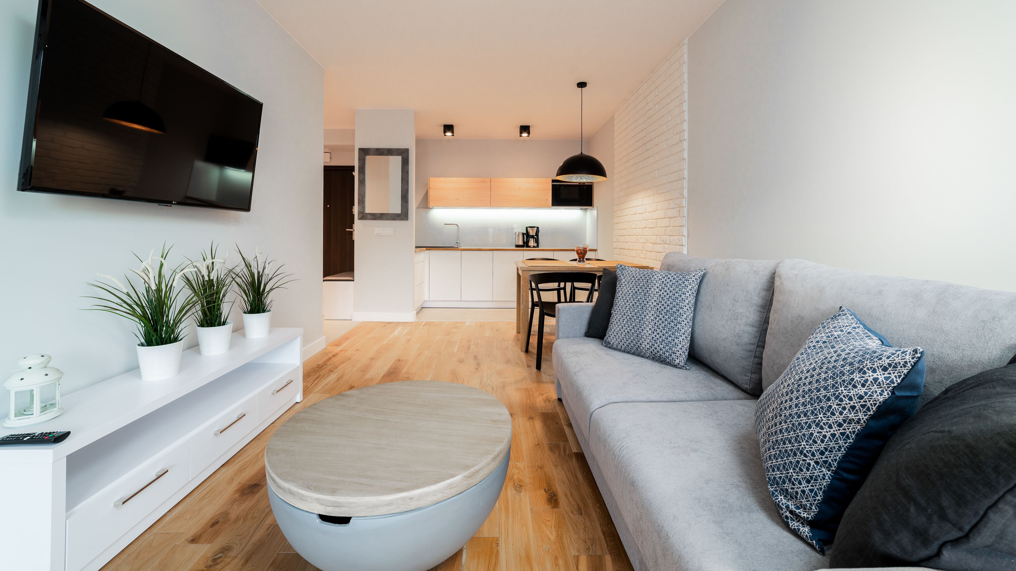 Una sala moderna con un sofá gris, un otoman redonda y un mueble blanco para entretenimiento con plantas. Se ve una cocina al fondo con una zona de comedor.