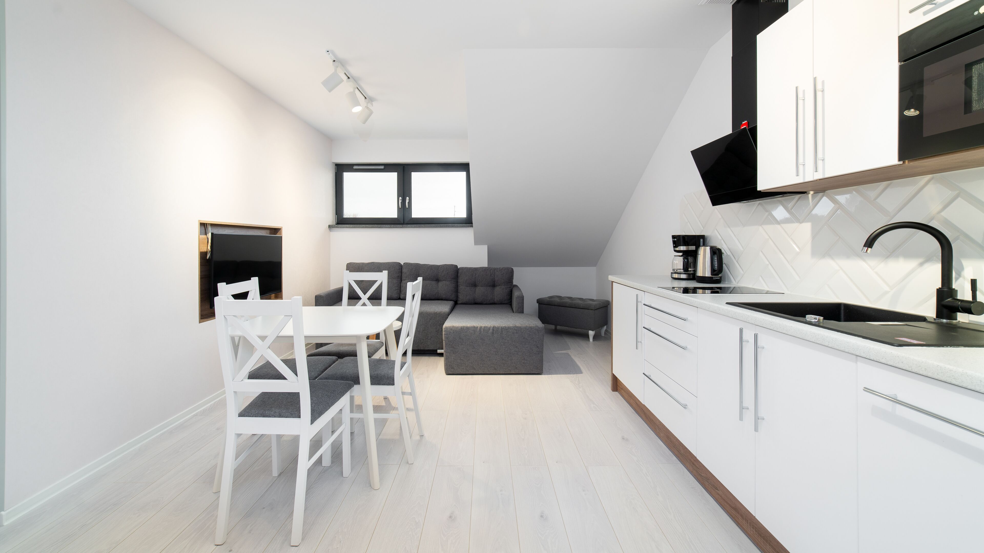A modern kitchen with white cabinets, a black faucet, and a built-in oven. There is a dining area with a white table and chairs, and a gray sofa.
