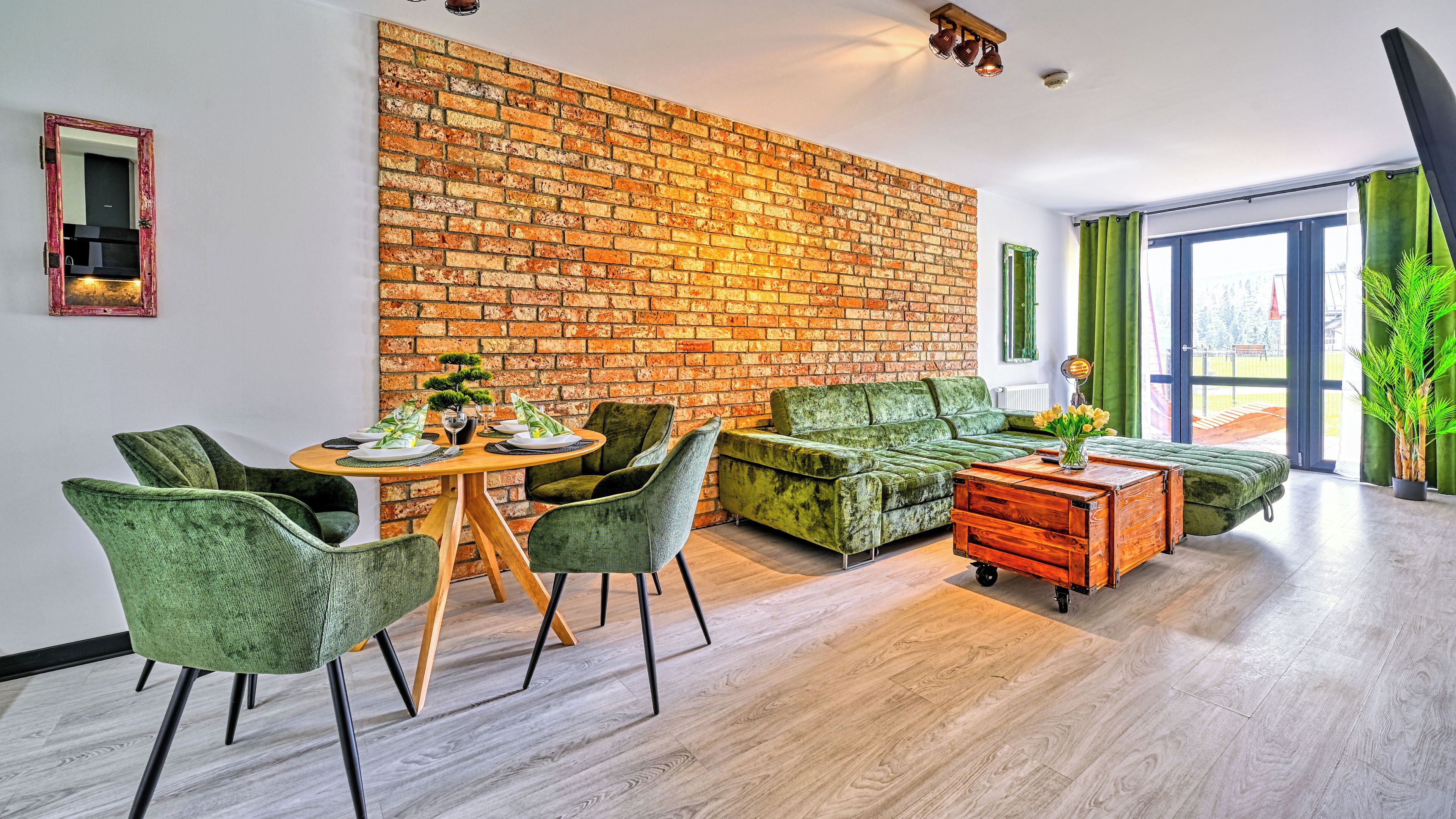 Ein Wohnzimmer mit Backsteinwand, grünen Möbeln, einem Esstisch mit Tellern und Blick durch eine Glastür.