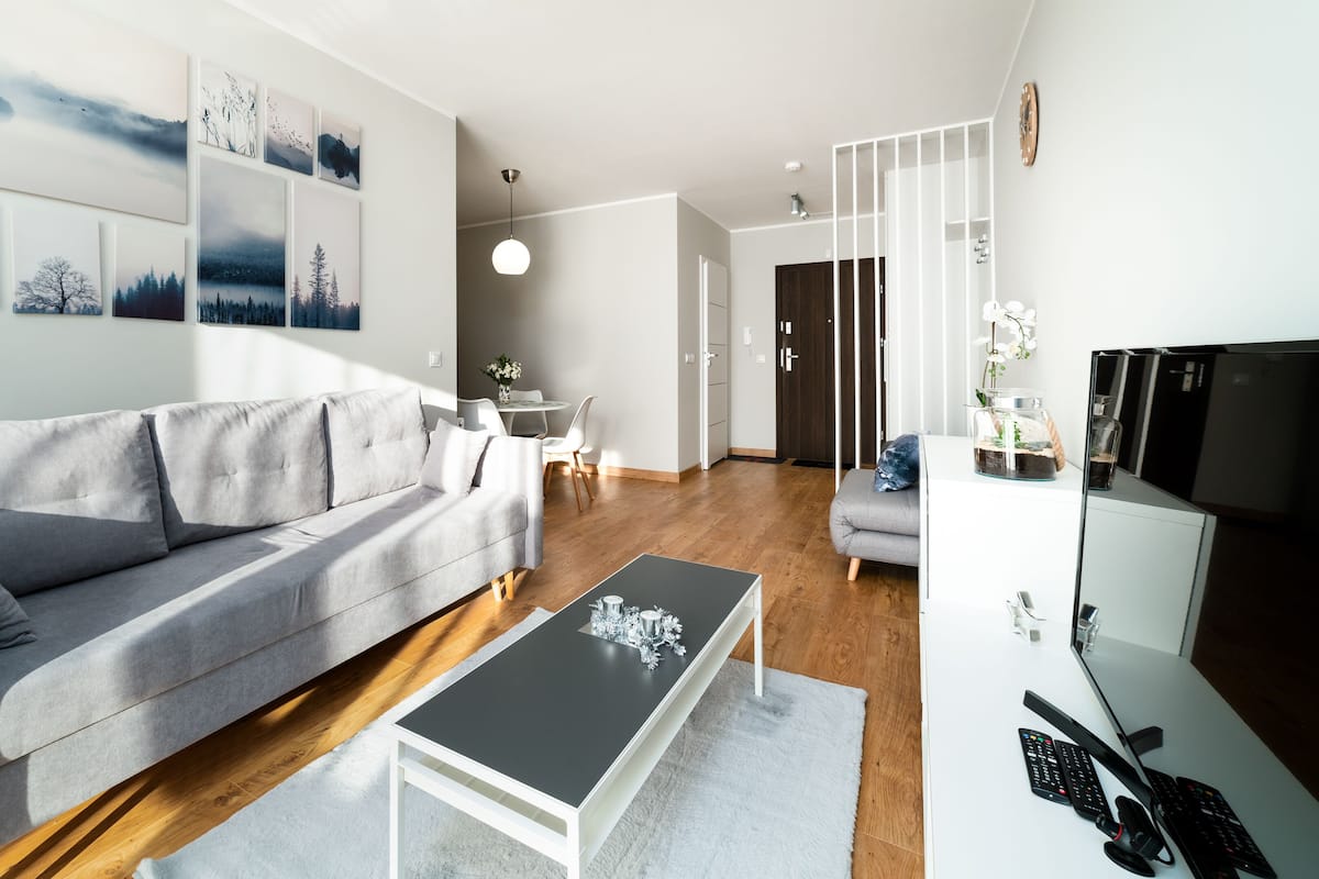 Una sala de estar moderna con un sofá gris, una mesa de centro y un mueble de TV con televisor de pantalla plana. Se observan obras de arte colgadas en la pared y un comedor al fondo.