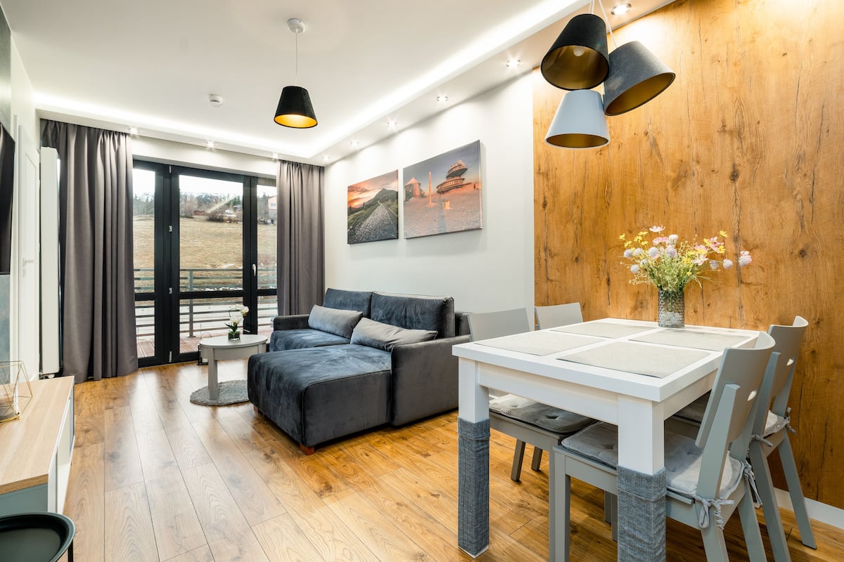 Un moderno salón con sofá, mesa de comedor y sillas. Hay dos lámparas colgantes, una pared de madera y una ventana con cortinas.