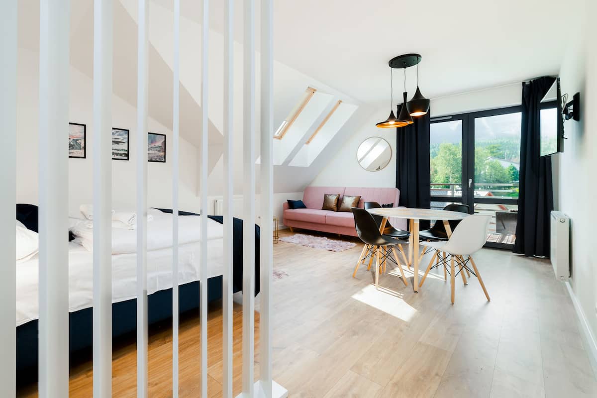 Un dormitorio moderno con cama, un sofá rosa, una mesa de comedor con sillas y un ventanal con vistas a zonas verdes.