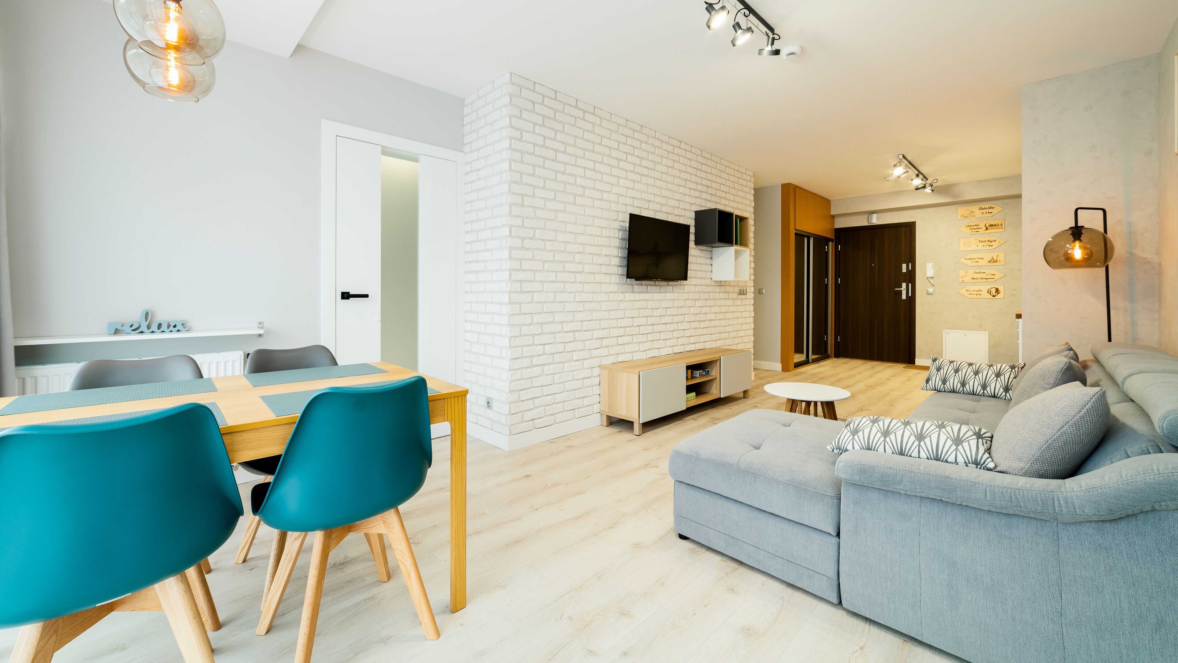 A modern living room with a grey sofa, teal chairs, a wooden dining table, and a white brick accent wall.