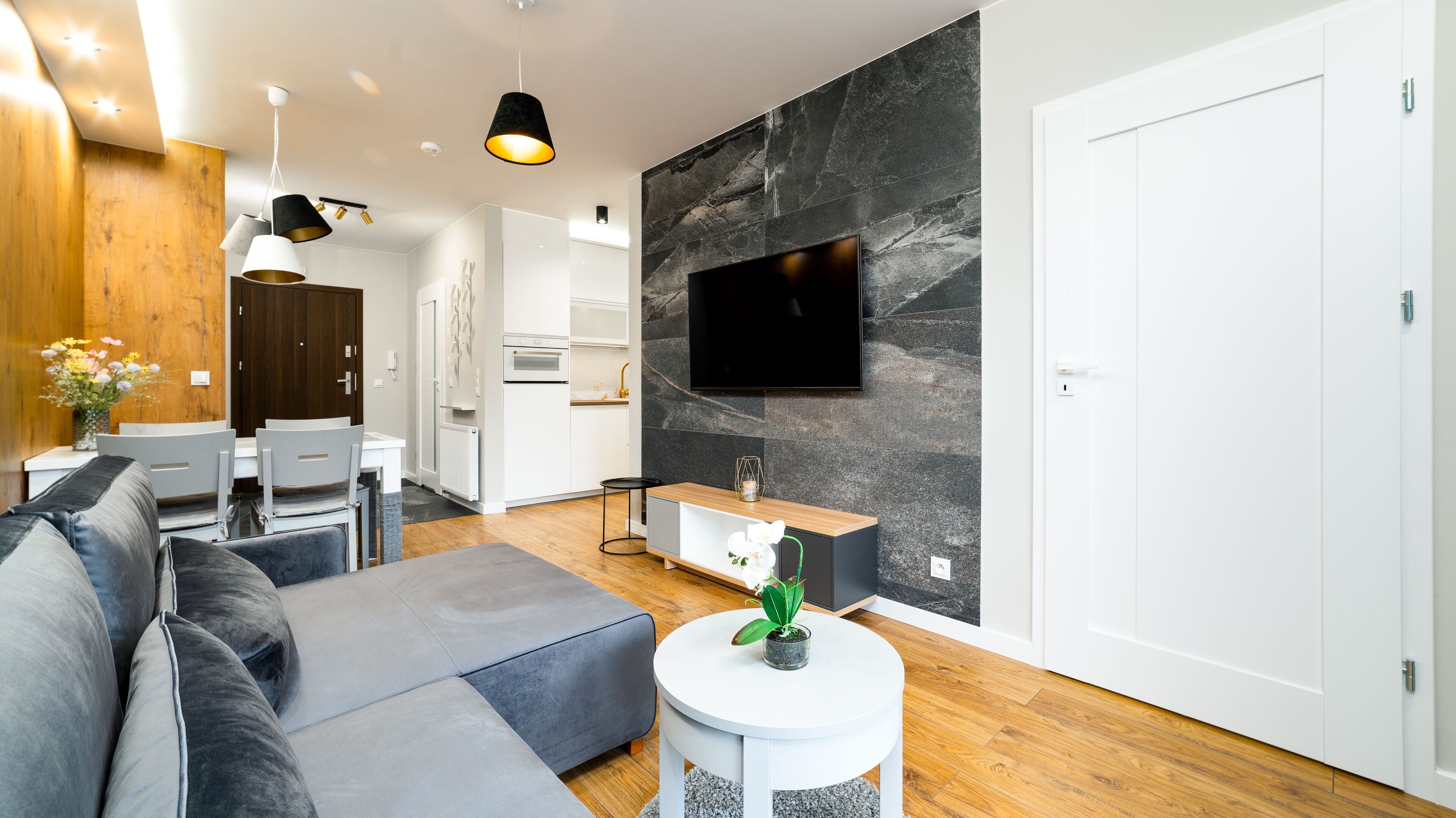 Una sala de estar moderna con un sofá gris, una pequeña mesa redonda con una planta, un televisor montado en una pared de mármol y una zona de comedor de madera.