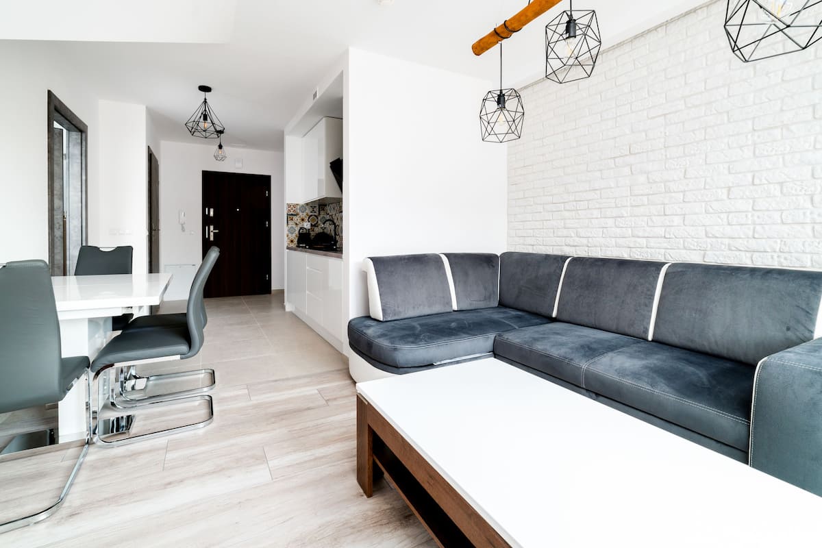 Una sala de estar moderna con un sofá seccional gris, una mesa de comedor blanca con sillas y una cocina al fondo.