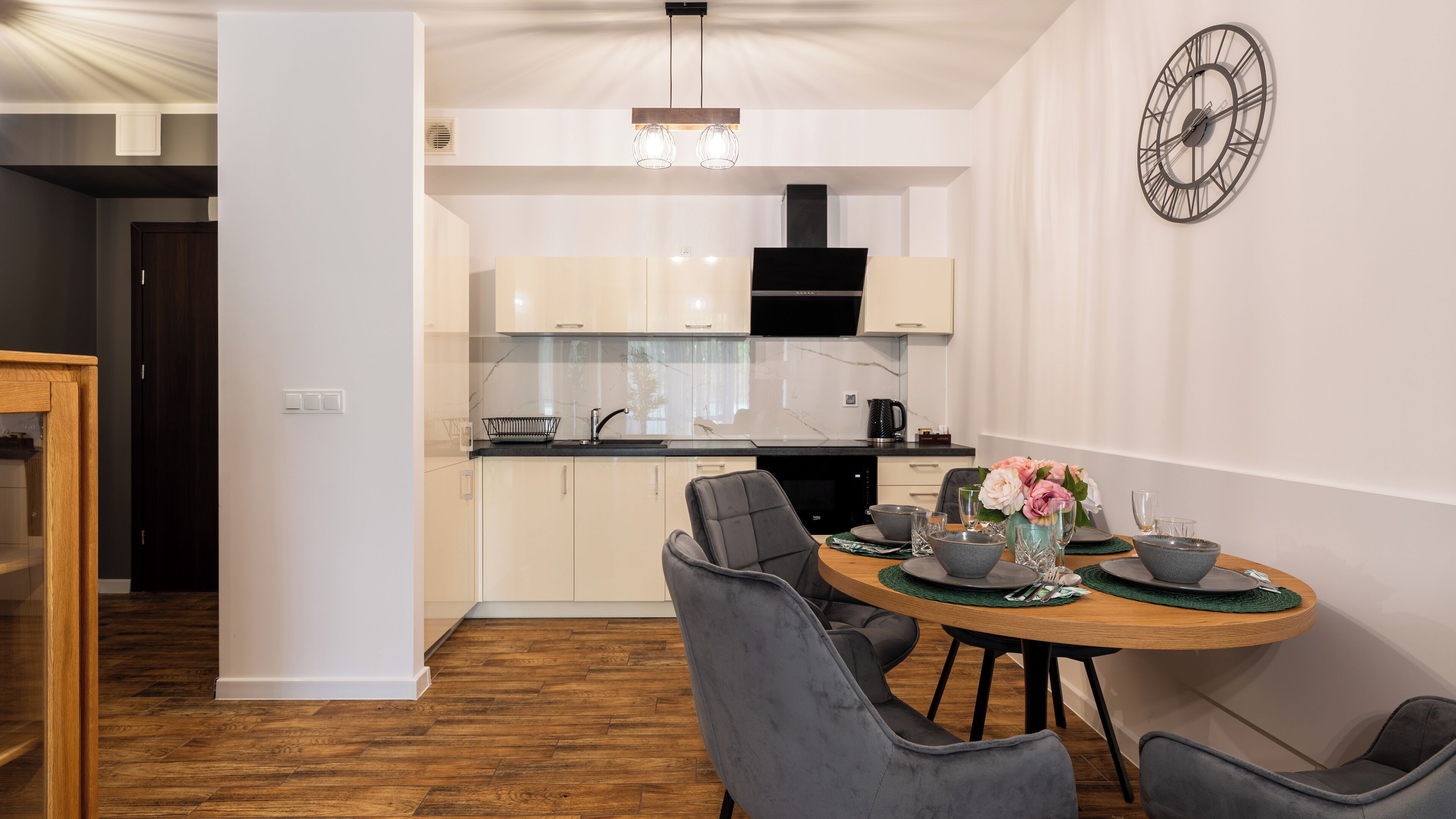 A modern kitchen with white cabinets, a black range hood, and a marble backsplash. There is a dining area with a round table, grey chairs, and a vase with flowers.