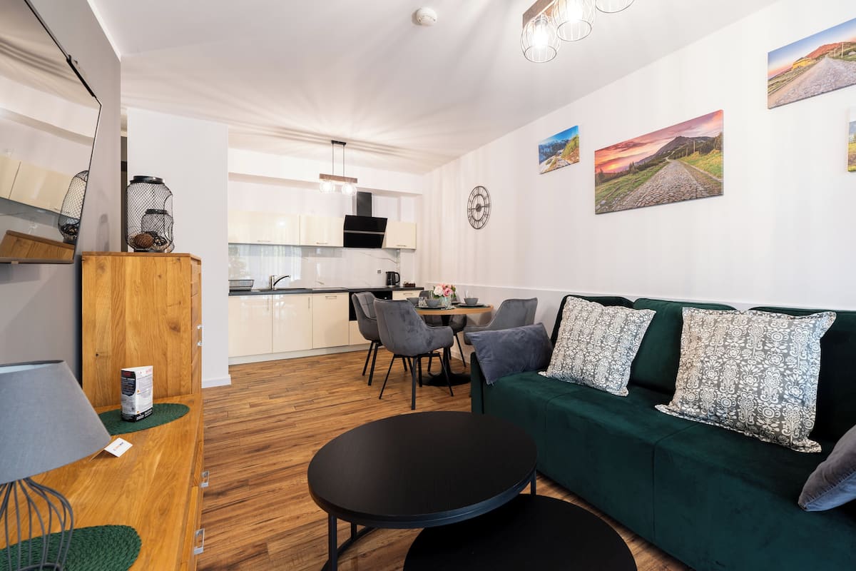 Una sala de estar moderna con un sofá verde, una mesa redonda negra y una cocina al fondo.