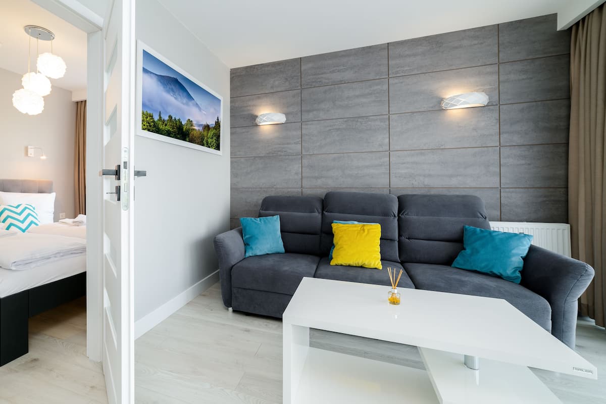 Habitación de hotel moderna con una cama, un sofá gris con cojines de colores, una mesa de centro blanca y un ventanal con vistas a la montaña.