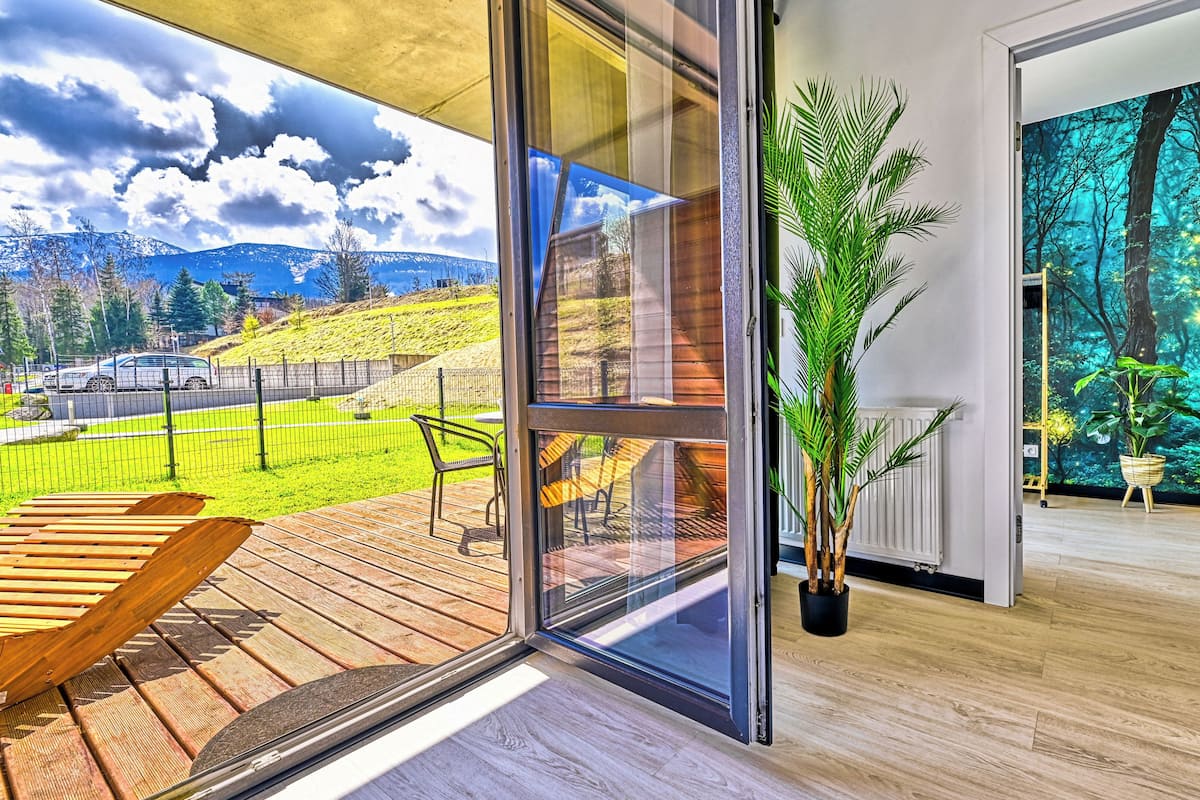Una habitación moderna con un amplio ventanal que da a una terraza exterior, una planta en maceta y un mural con motivos forestales.