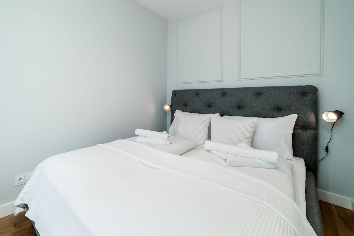Una cama bien hecha con sábanas blancas, un cabecero tapizado en gris y dos lámparas de lectura fijadas a la pared.