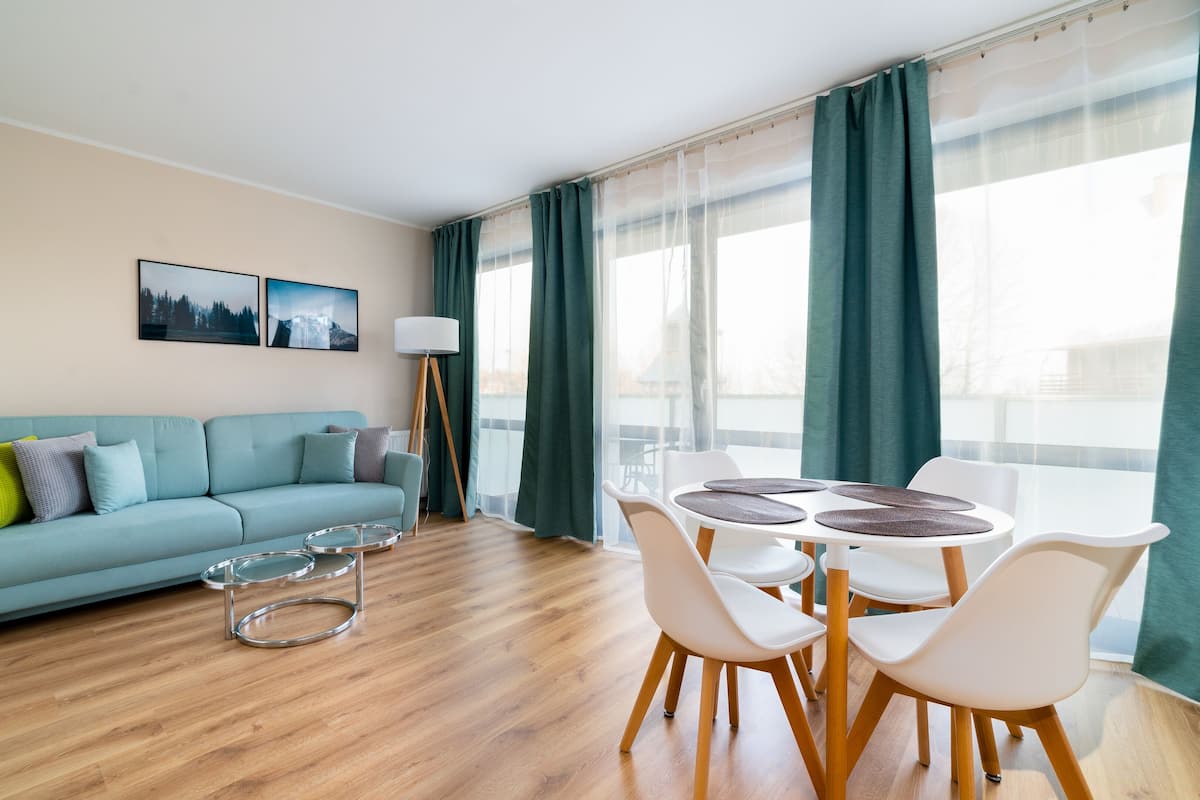 Una sala de estar moderna con un sofá azul claro, una mesa de comedor con sillas blancas y un ventanal con cortinas verdes.