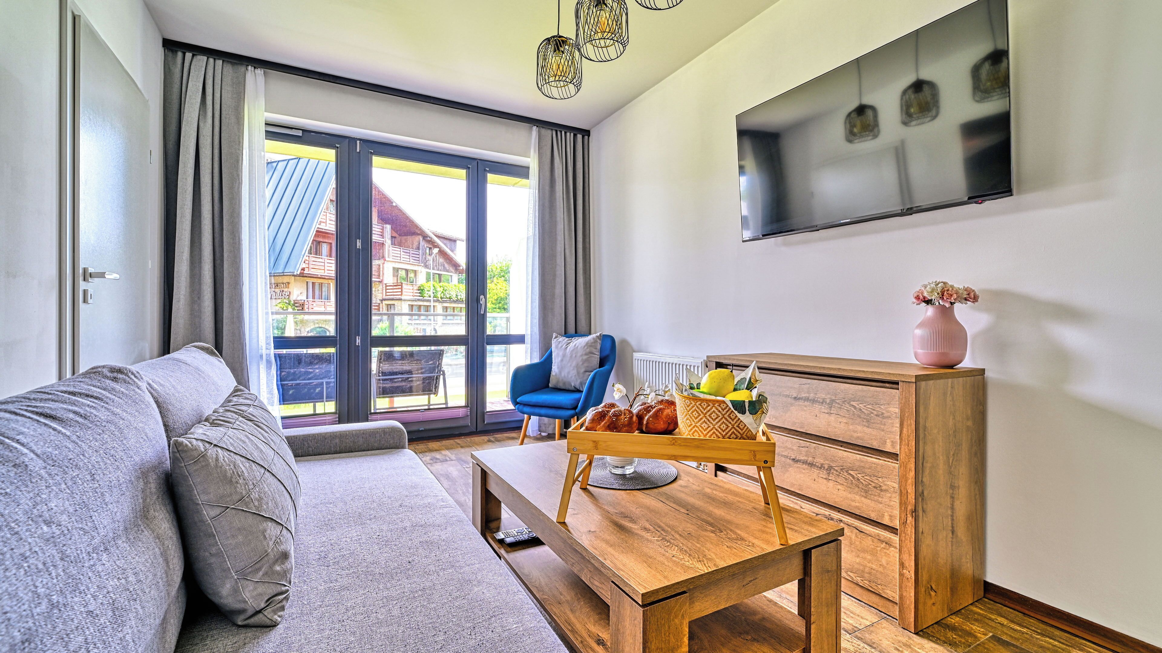 A modern living room with a sofa, a blue armchair, a wooden coffee table with fruits and flowers, and a TV mounted on the wall.