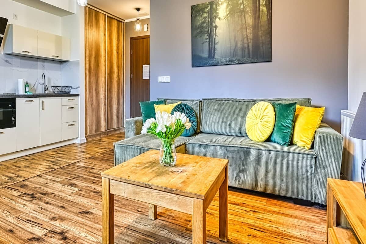 Una sala de estar con un sofá, una mesa de centro con un jarrón de flores, una lámpara y una cocina al fondo.