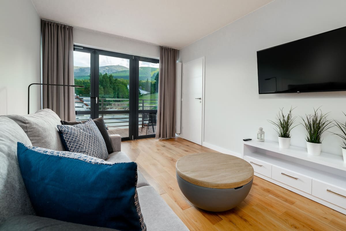 Una sala de estar moderna con sofá, un reposapiés redondo, televisor montado en la pared y un balcón con vistas a zonas verdes y colinas.