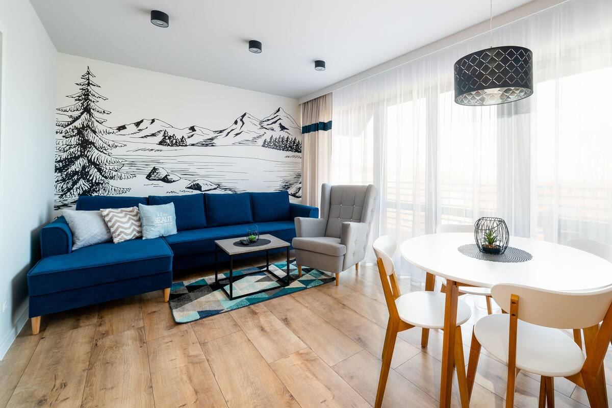 Una sala de estar moderna con un sofá azul, una mesa de comedor con sillas, una alfombra estampada y un mural en la pared.