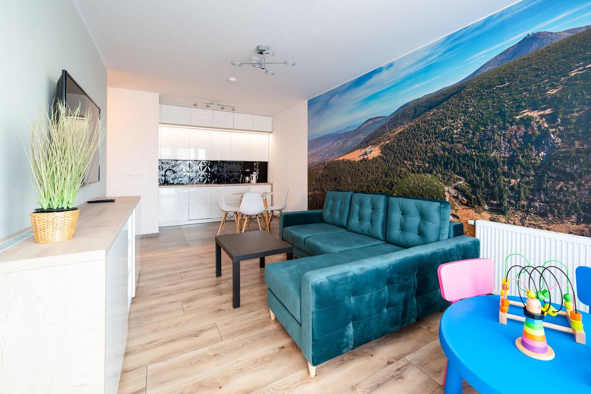 Una sala moderna con un sofá azul turquesa, una mesita y un juguete colorido. Se ve una cocina en el fondo, con armarios blancos y un salpicadero negro.