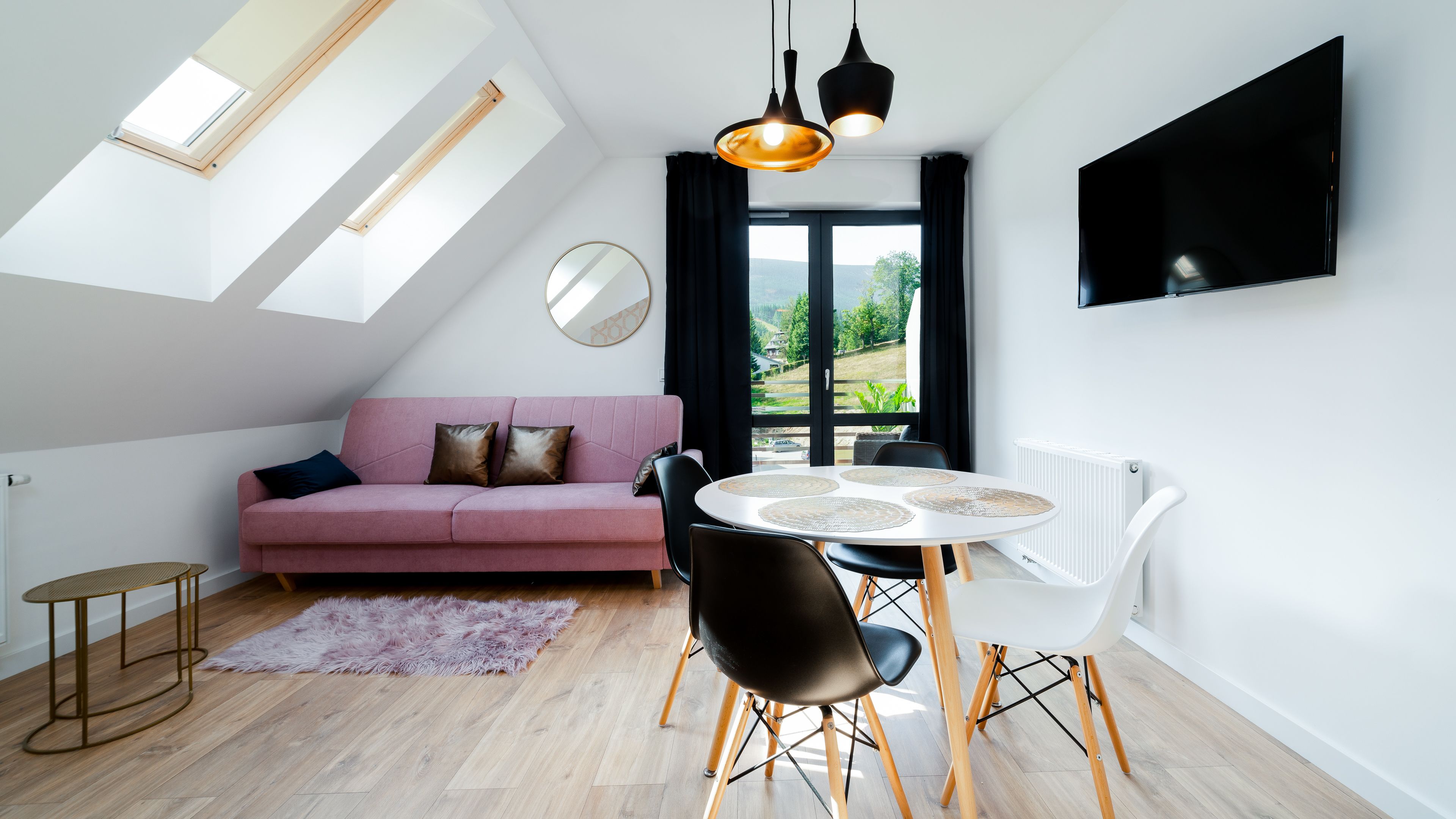 A modern living room with a pink sofa, a round dining table with chairs, a large window with a view of greenery, and a flat-screen TV mounted on the wall.