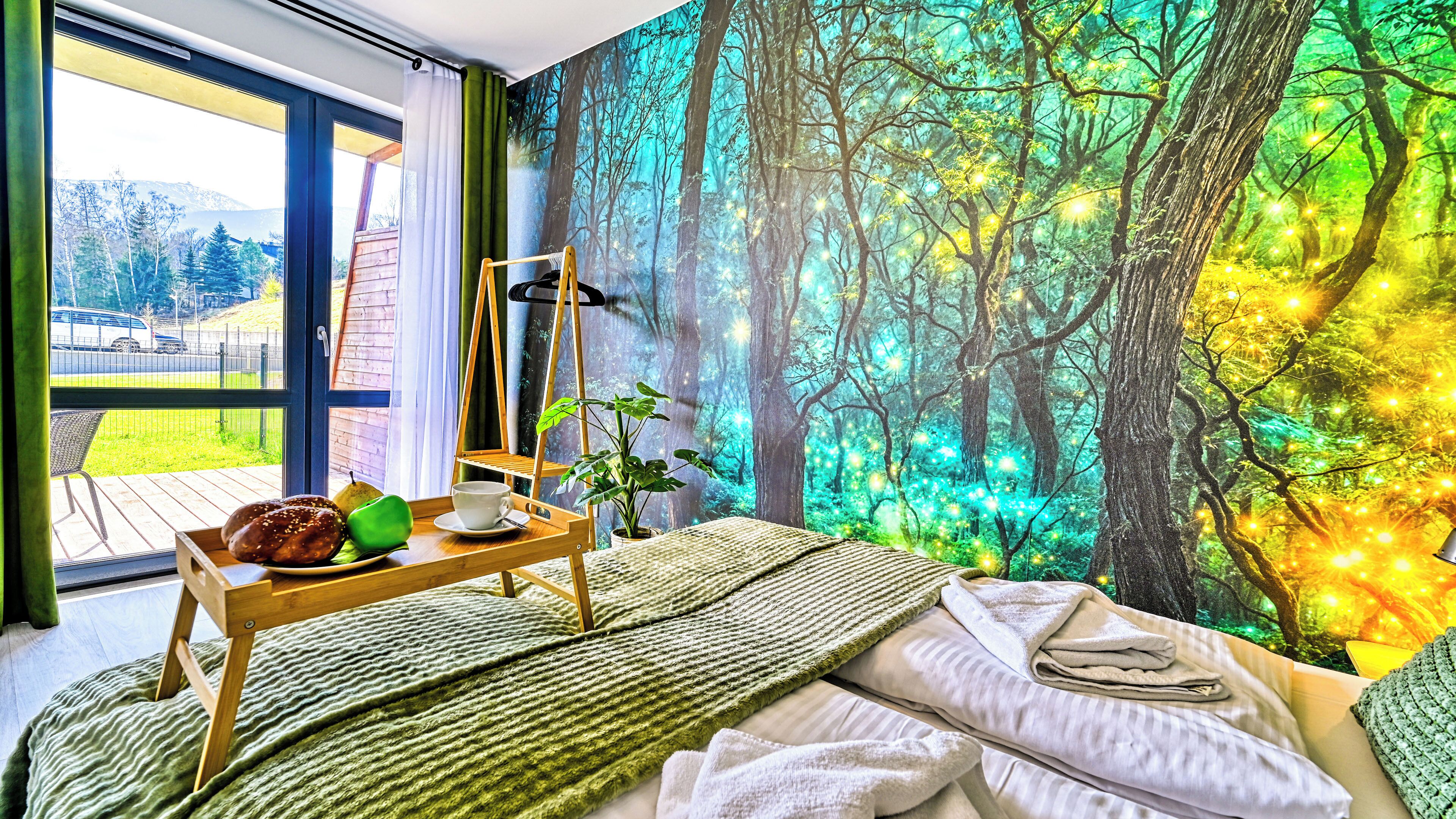 A room with a bed, a wooden side table with breakfast items, a window with a view of a forest, and a wall mural of a forest.