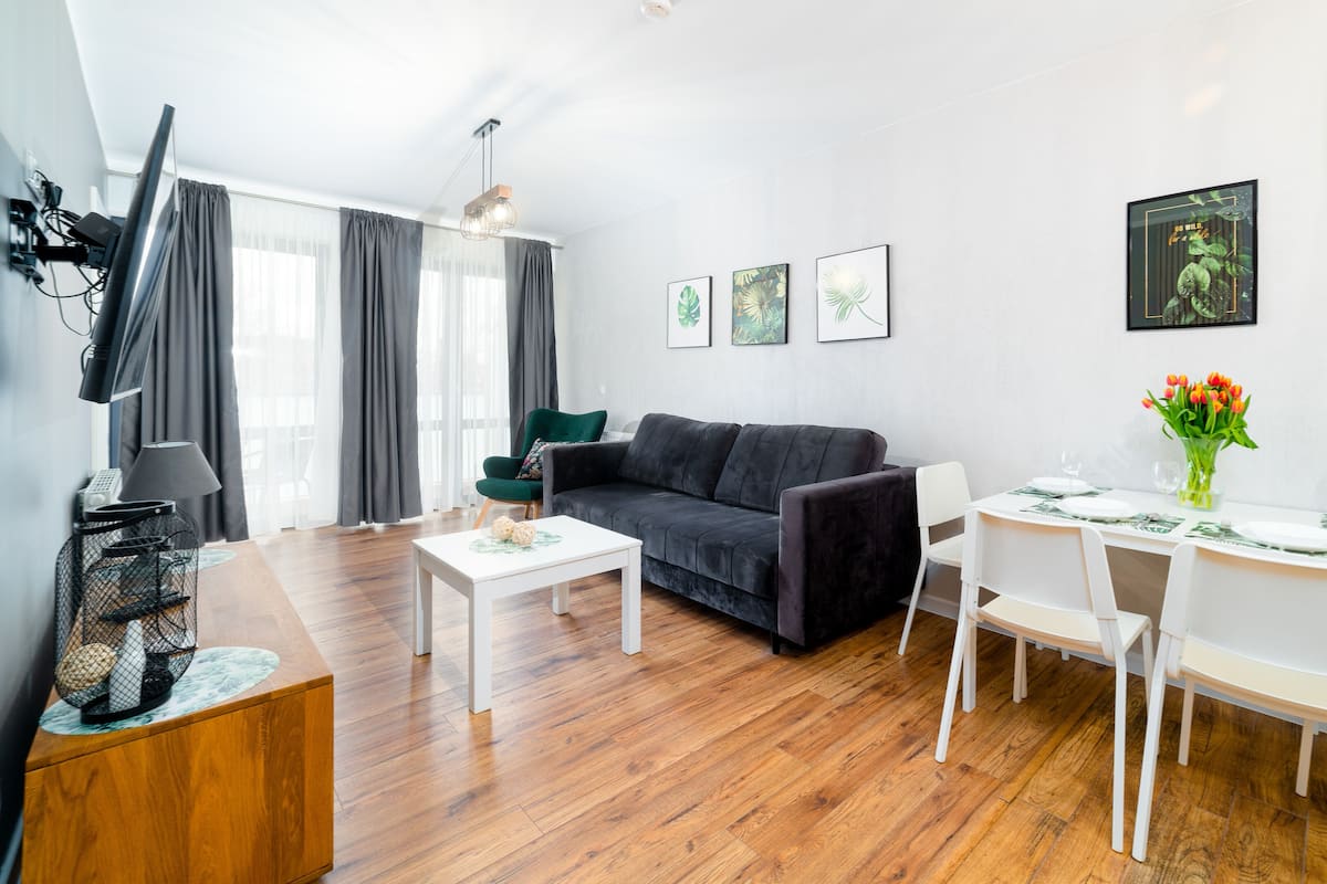 Una sala de estar moderna con un sofá gris oscuro, una mesa de comedor blanca y sillas. Hay cuadros en la pared, un televisor de pantalla plana montado y una pequeña mesita de noche con una lámpara.