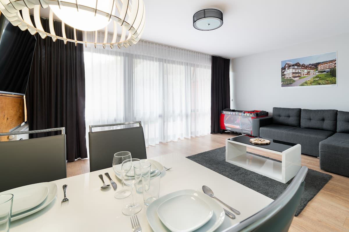 Una sala moderna con un sofá gris, una mesa de comedor con platos y vasos, un televisor y un ventanal con cortinas de gasa.
