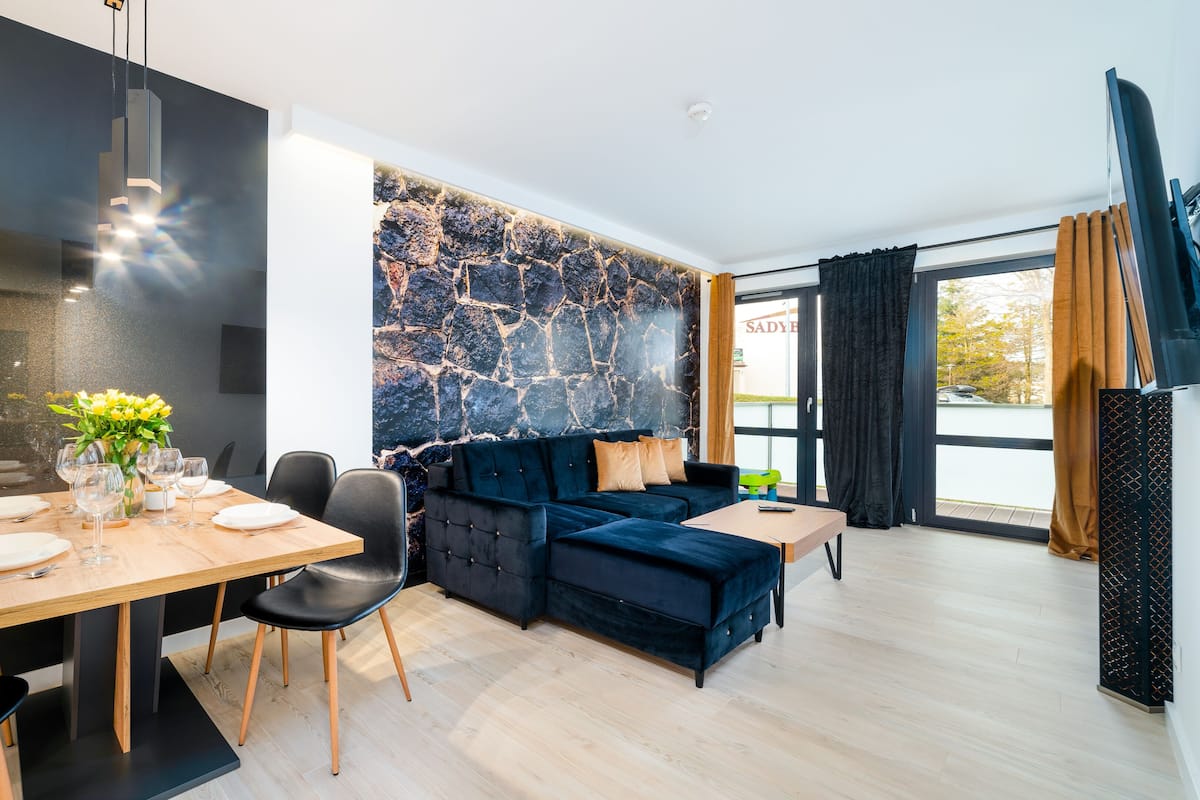 Una sala de estar moderna con comedor, que incluye un sofá azul oscuro, una mesa de comedor de madera para cuatro personas y una pared de piedra.