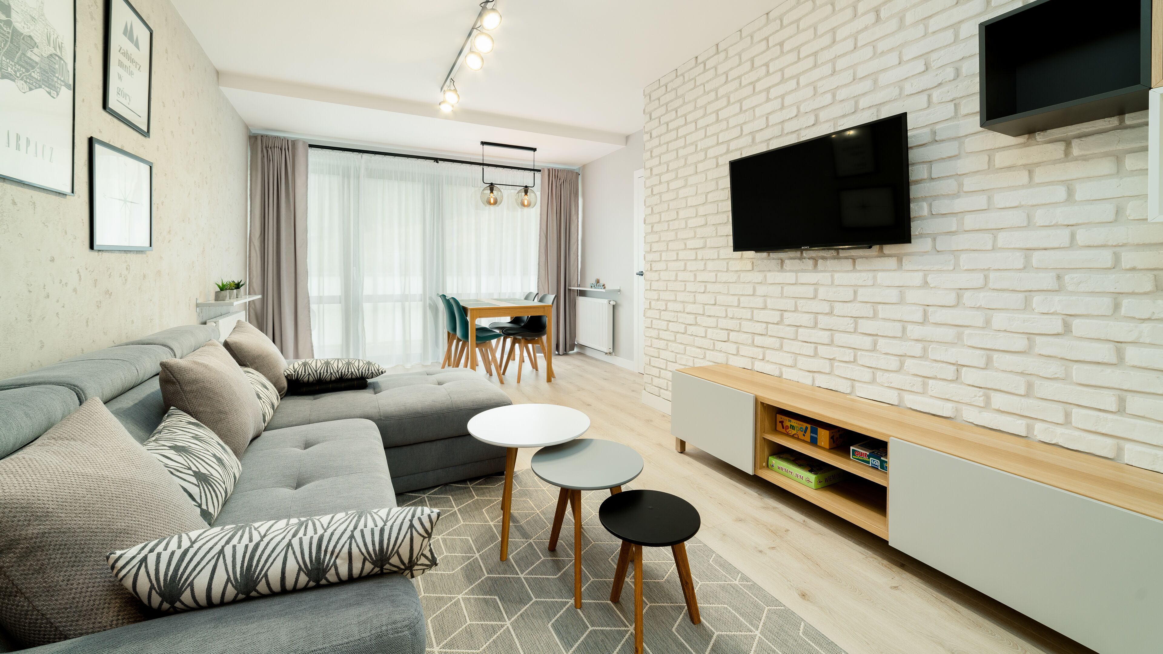 Un salon moderne avec un canapé gris, un téléviseur à écran plat fixé sur un mur en briques blanches et une petite table d’appoint en bois.