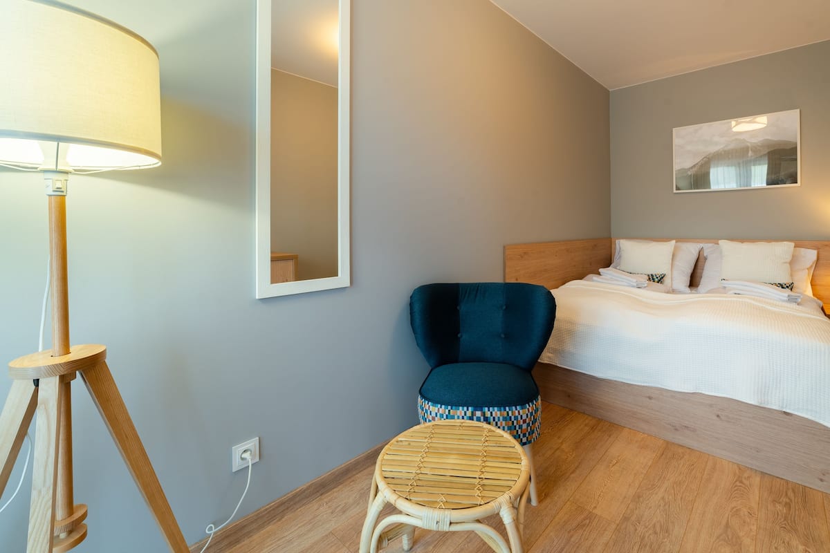 Habitación de hotel con cama, una silla, un taburete pequeño, mesita de noche con lámpara, espejo y un cuadro en la pared.