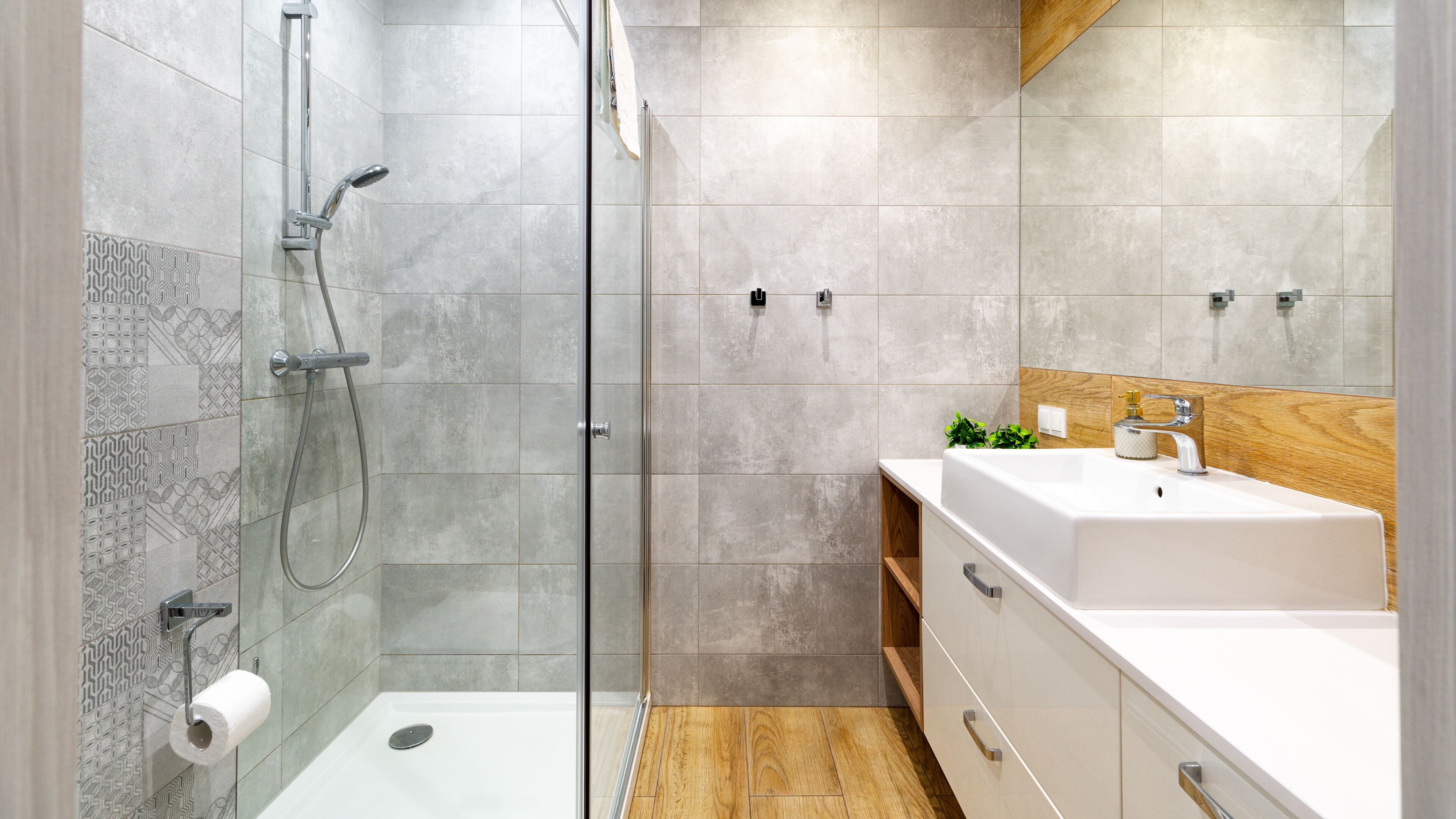 A modern bathroom with a glass shower enclosure, a white sink with a wooden countertop, and patterned tiles on the walls.