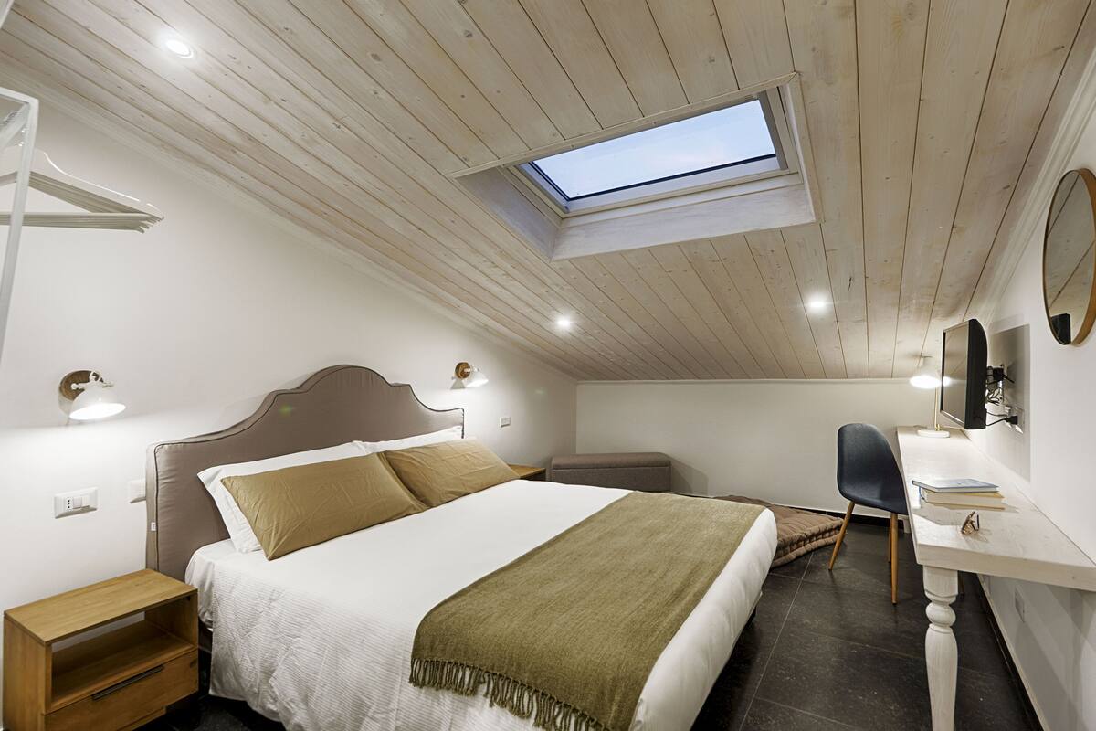 A bedroom with a skylight, a bed with a headboard, bedside tables, a desk with a chair, and a mirror on the wall.