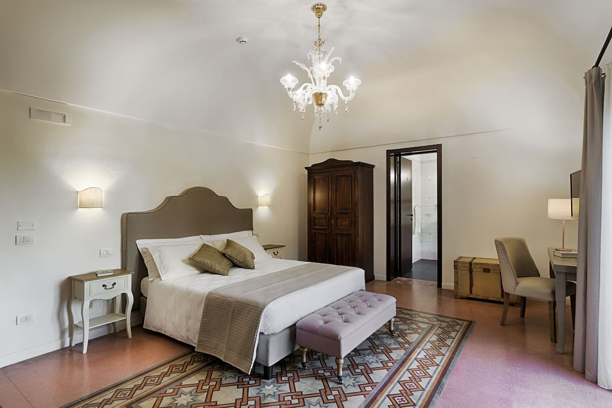 A bedroom with a large bed, a wooden wardrobe, a bench, a nightstand, and a chandelier.
