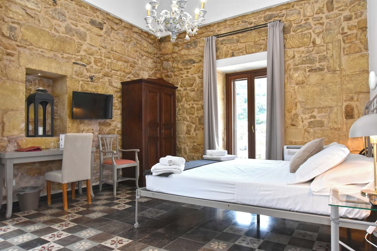 A bedroom with a stone wall, a bed, a desk, a chair, a TV, and a chandelier.
