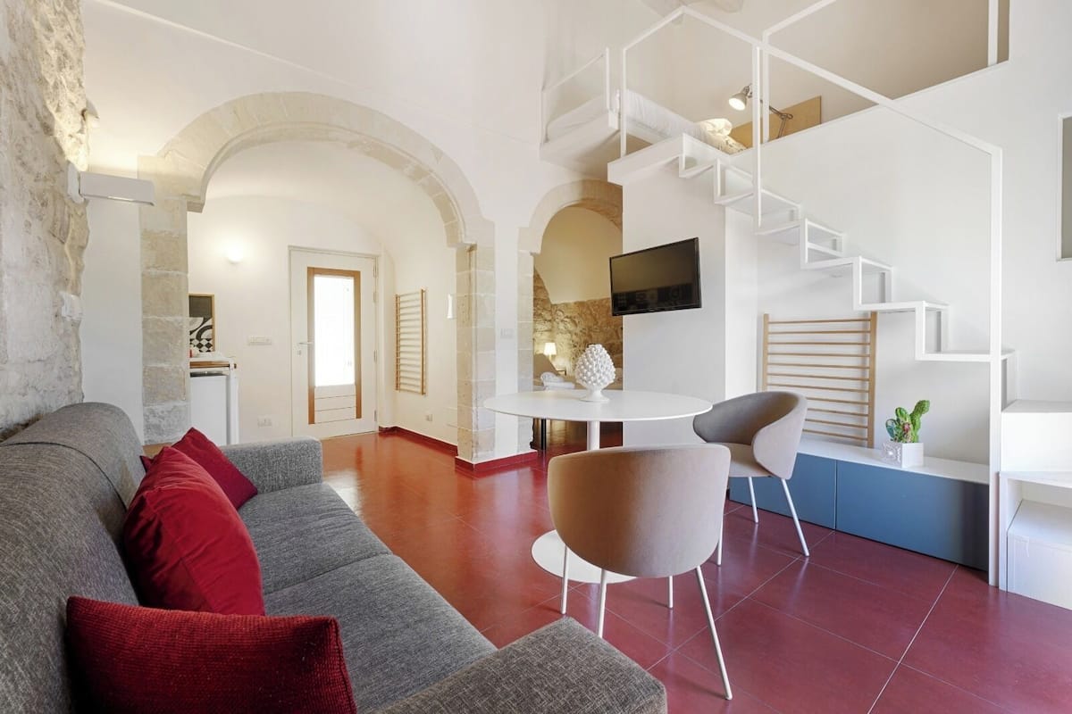 A modern living room with a grey sofa, red cushions, a round white table, and two chairs. There is a TV mounted on the wall, a staircase with white railings, and a stone wall.