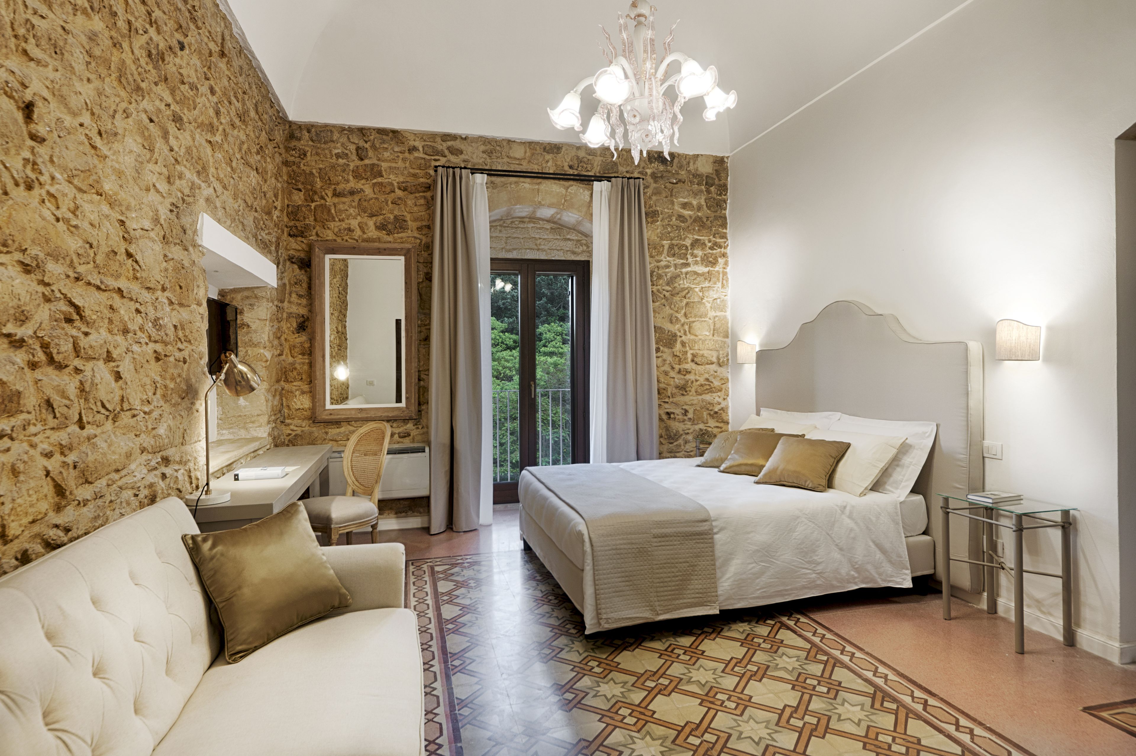 Une chambre avec un mur en pierre, un lit avec une tête de lit, une table de chevet, un bureau, une chaise et un lustre.