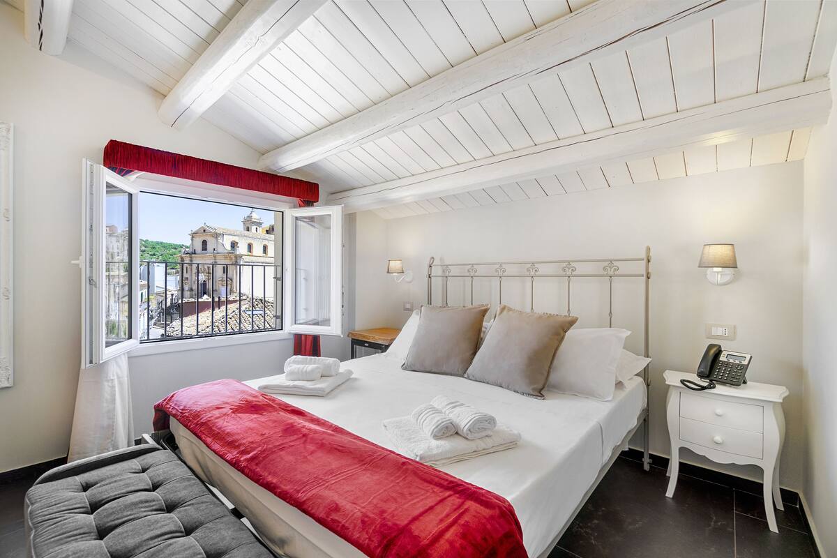 A bedroom with a bed, bedside table, lamp, and a window with a view of a building.