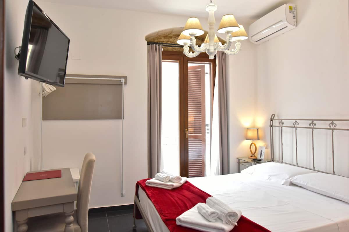 A hotel room with a bed, a desk, a chair, a TV, a chandelier, and an air conditioning unit.