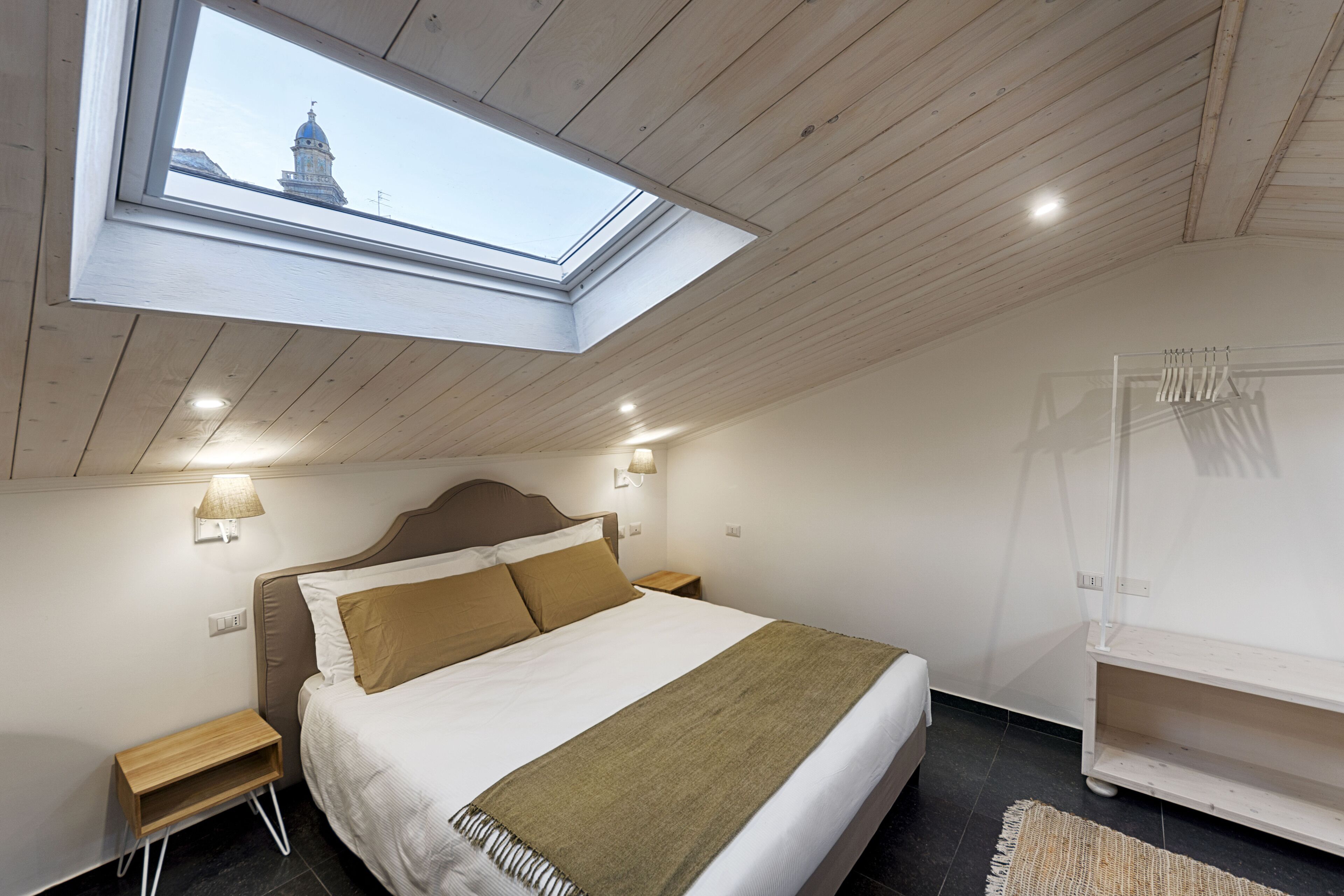 Ein Schlafzimmer mit Oberlicht, einem Bett mit Kopfteil, Nachttischen, einem Wandregal und Blick auf ein Gebäude durch das Oberlicht.