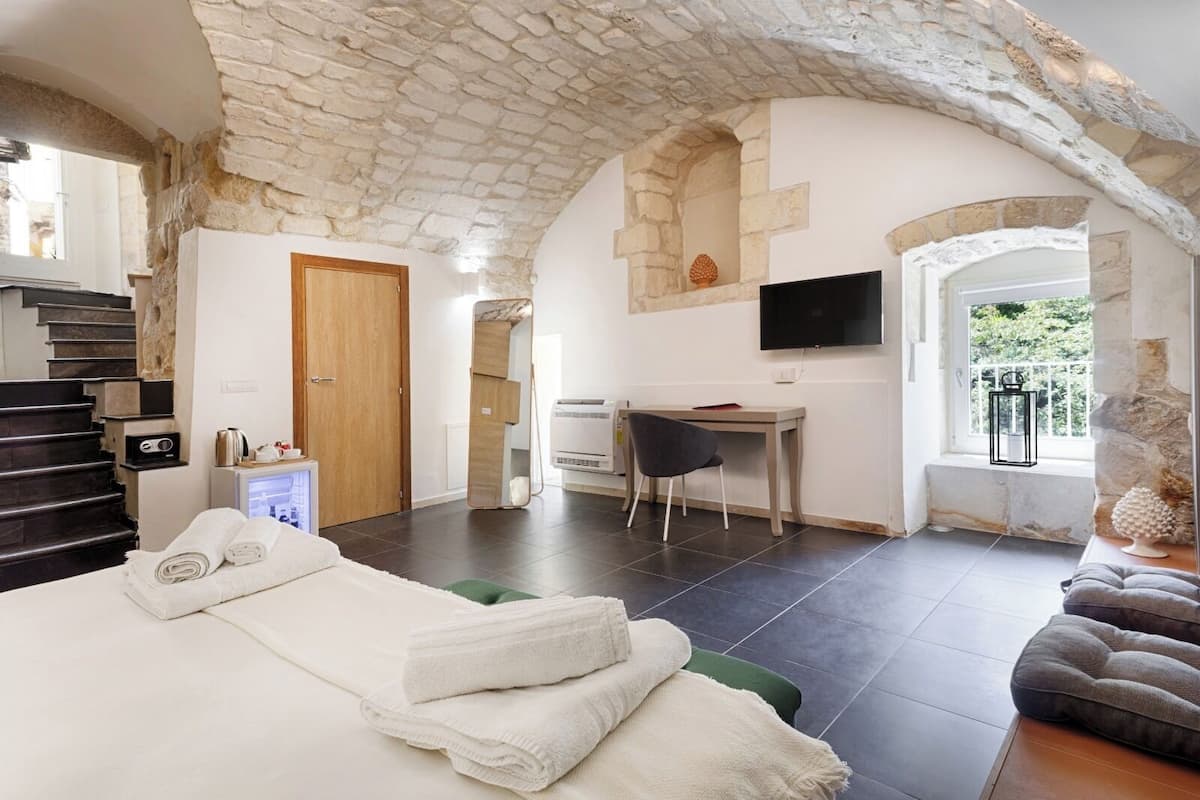 A hotel room with a stone ceiling, a bed with towels, a desk with a chair, a TV, and a window with a view of greenery.