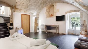A hotel room with a stone ceiling, a bed with towels, a desk with a chair, a TV, and a window with a view of greenery.