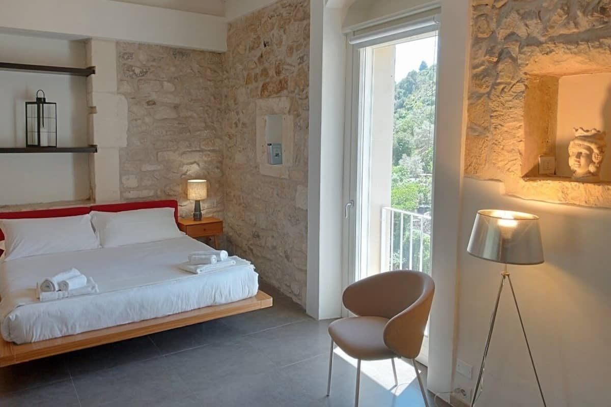 A bedroom with a stone wall, a bed with white linens, a bedside table with a lamp, a chair, and a view of greenery outside.