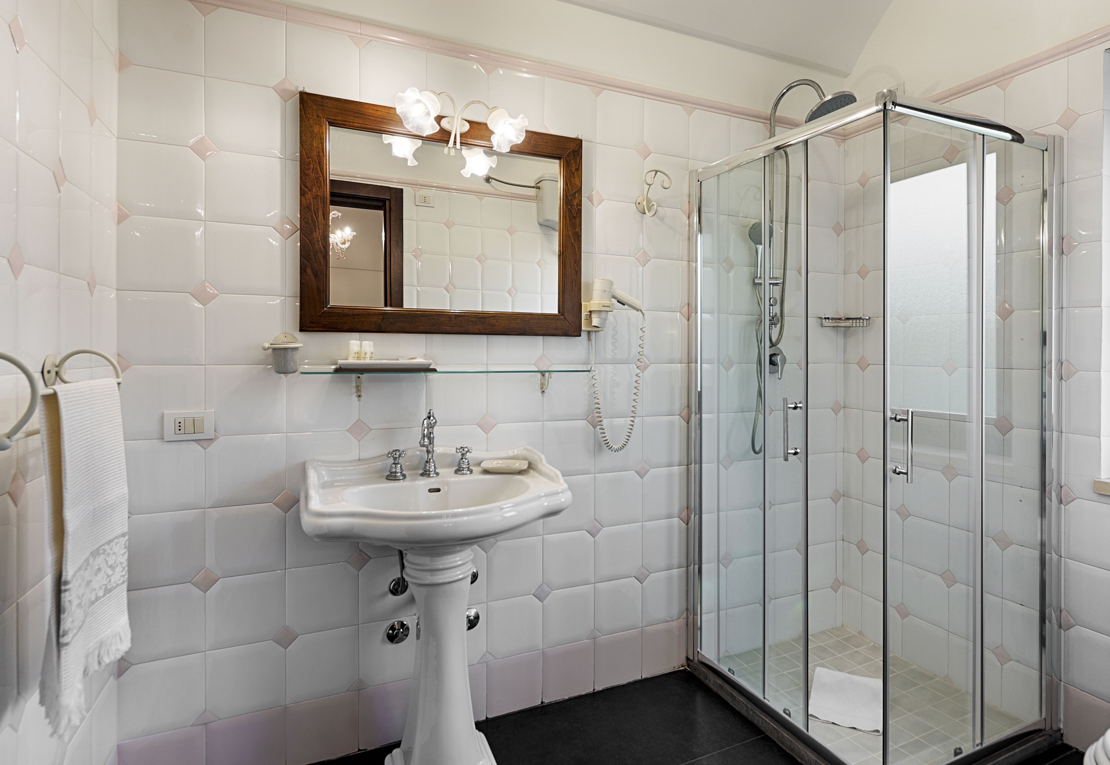 Een badkamer met een wastafel op een voetstuk, een spiegel met houten lijst, een douche met glazen wanden en een handdoekhouder.
