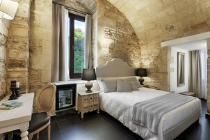 A bedroom with a stone wall, a bed with white bedding, a desk with a lamp, and a window with curtains.
