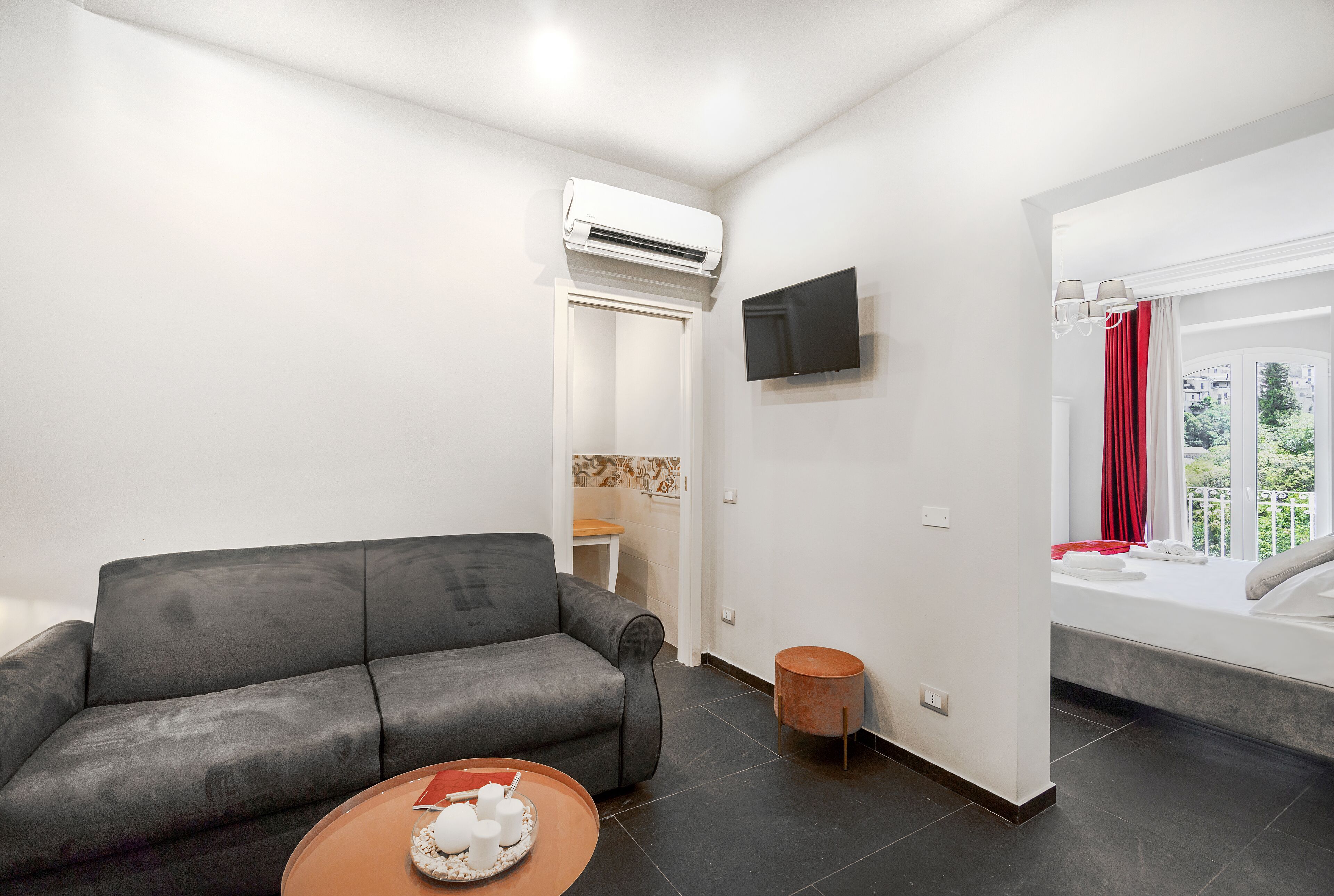 Ein modernes Wohnzimmer mit einem grauen Sofa, einem kleinen runden Tisch mit einer Auflage und verschiedenen Gegenständen, einem flachbildfernseher an der Wand und einem Fenster mit rotem Vorhang.