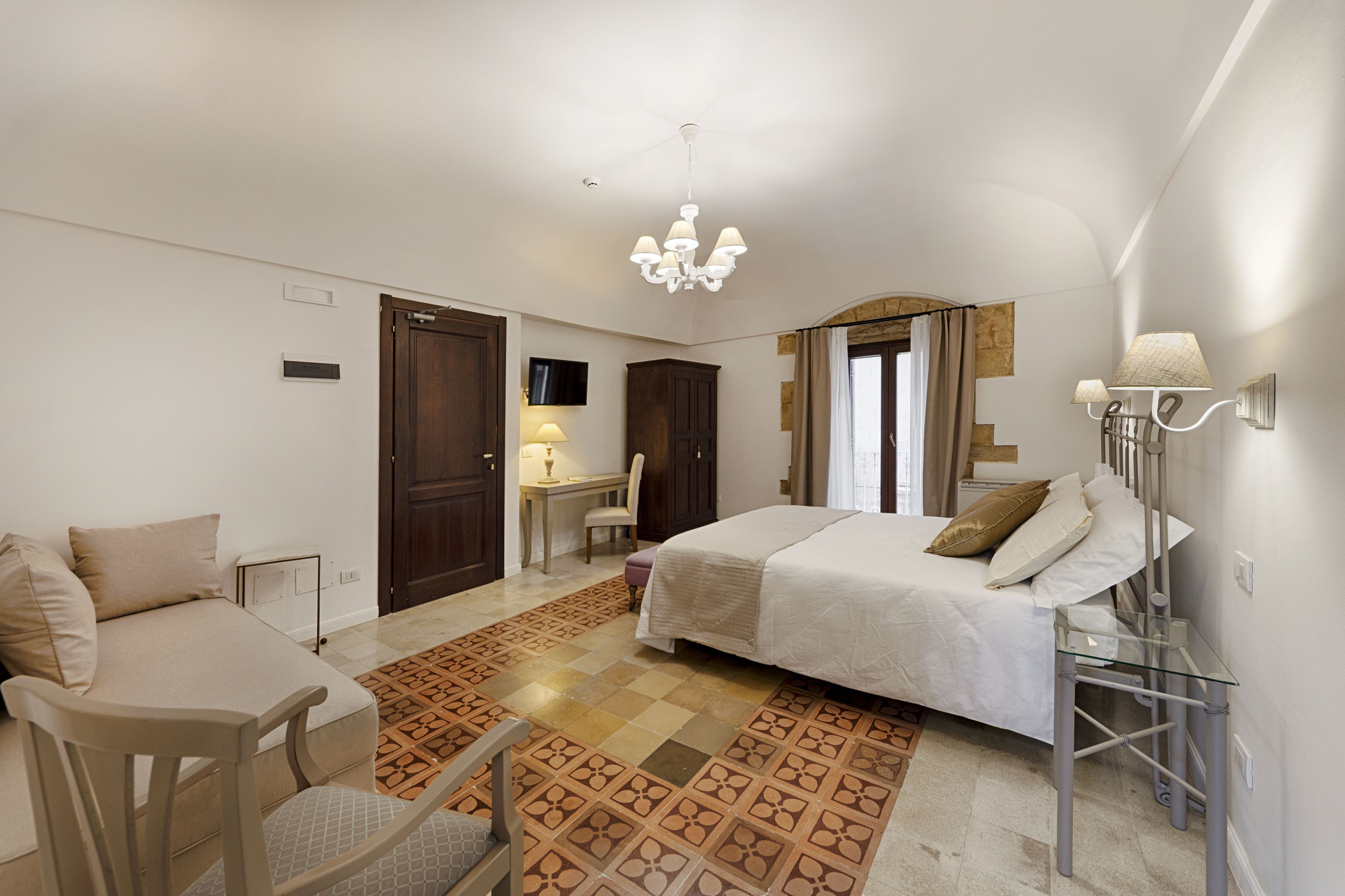 A hotel room with a bed, a desk, a chair, a TV, and a chandelier.