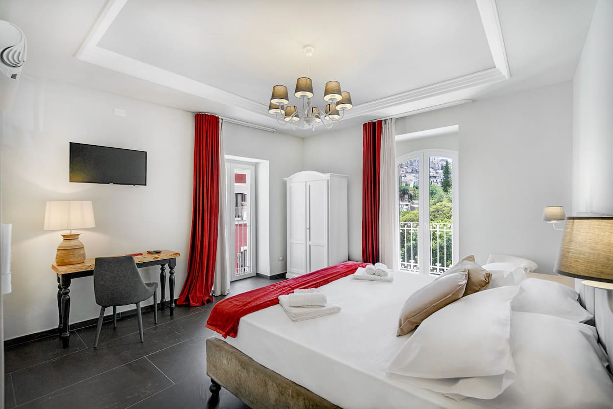 A hotel room with a bed, a desk with a lamp, a chair, a wardrobe, and a view of the outdoors.