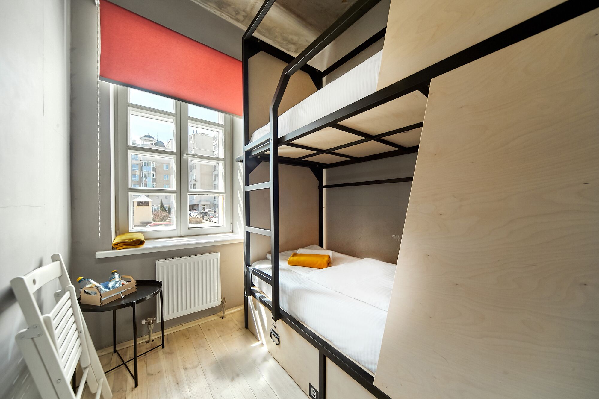 Une chambre avec un lit superposé, une fenêtre, un radiateur et une petite table avec des bouteilles.