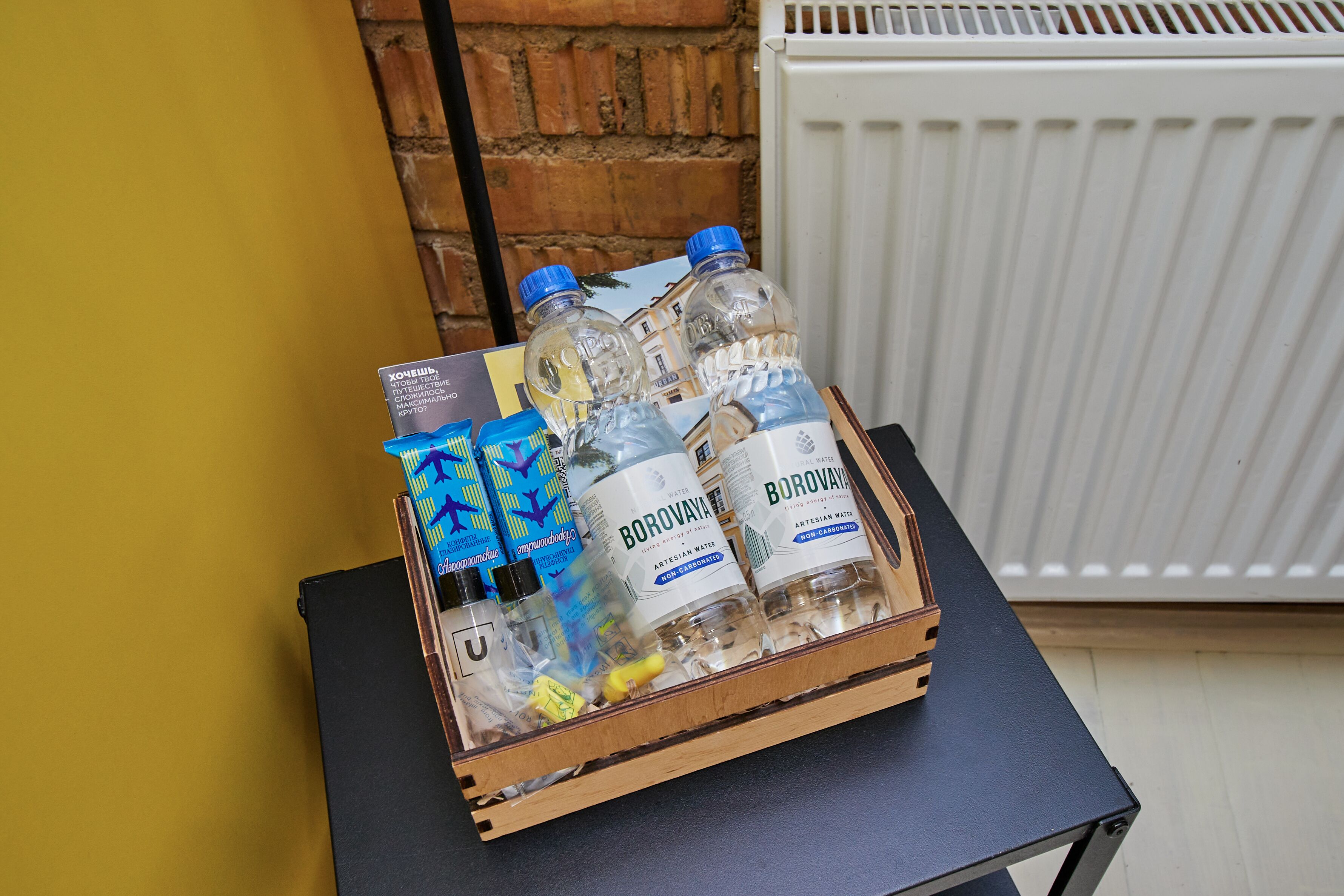 A wooden box containing bottled water and a small bottle of hand sanitizer on a black table against a yellow wall and a brick wall.